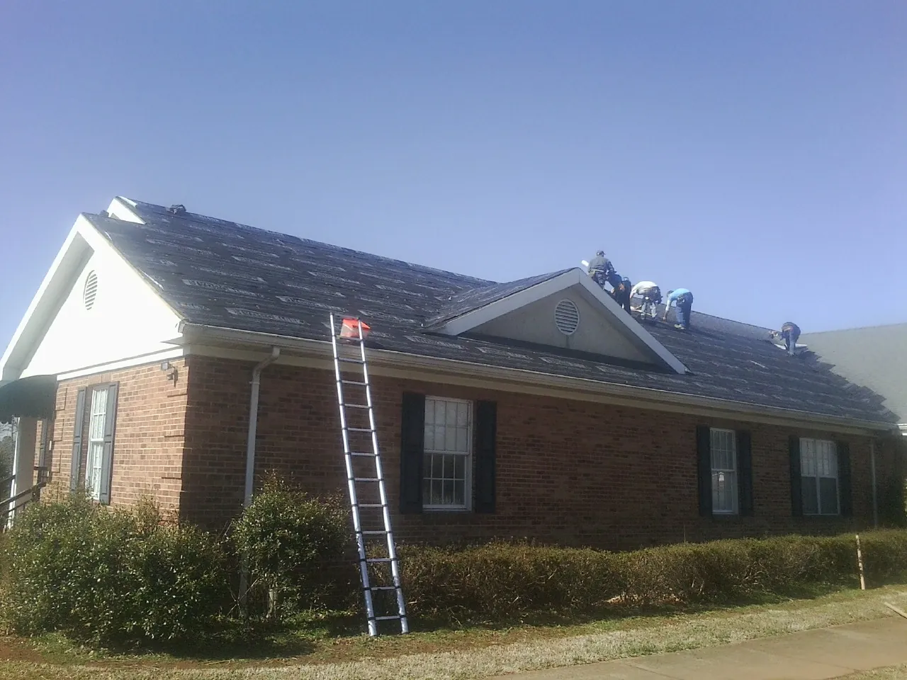 Crew tearing shingles off a brick ranch roof with a ladder set — Emergency Roof Repair work by our crew in Whitney Point