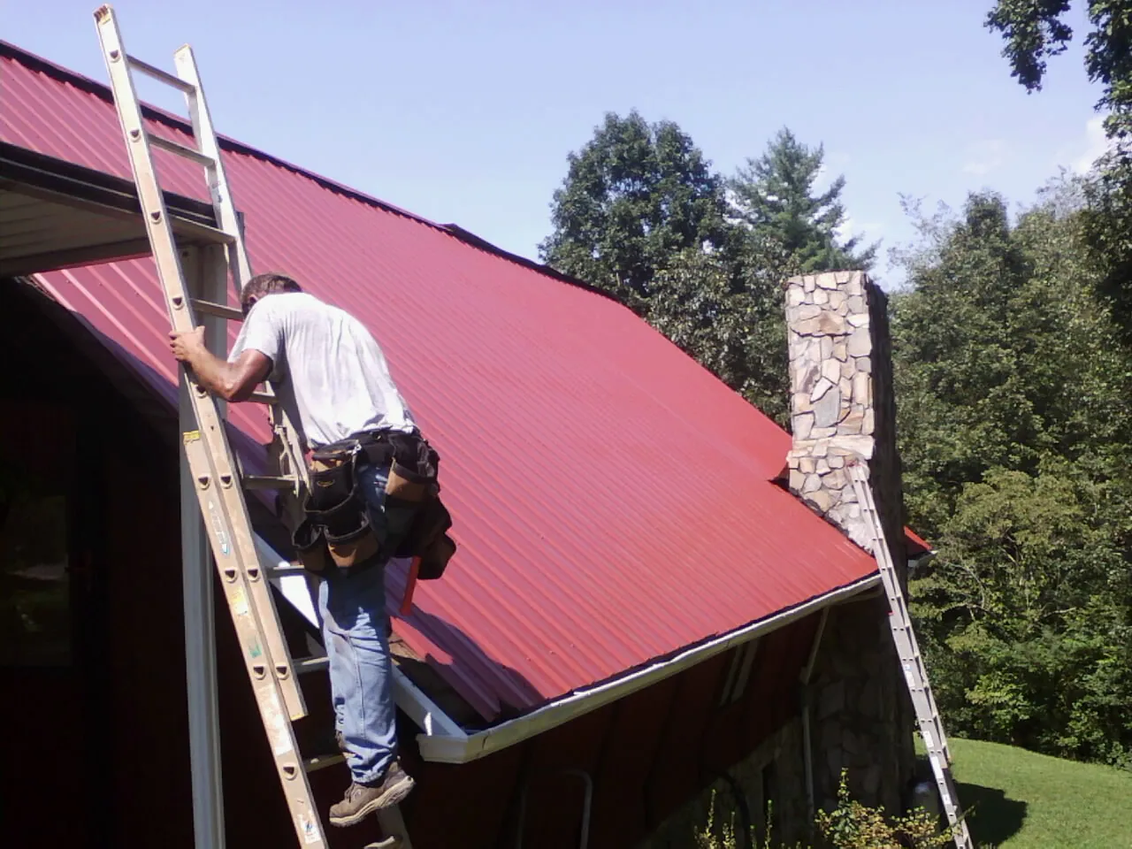 Roofer on a ladder beside a red metal roof and stone chimney — Soffit & Fascia Repair by Roofing Brothers in Whitney Point